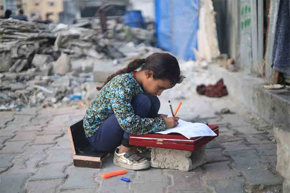 한 어린이가 길에서 공책에 무언가 쓰고 있다. 벽돌 위에 납작한 나무 상자를 얹어 책상으로 삼았고 합판으로 만든 간이 의자에 앉아 있다. 의자도 책상도 모두 낮아서 바닥에 쪼그리고 앉은 자세다. 뒤로는 무너진 건물 잔해가 보인다.